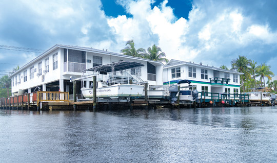 Photograph of a waterfront building complex with boats docked alongside a wooden pier under a partly cloudy sky.