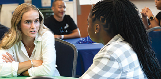 Photograph of two women engaged in a focused conversation at a table during a group event or meeting.