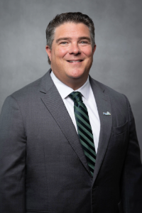 Brian Barrio, FGCU's director of intercollegiate athletics, poses for a photo. He is wearing a gray blazer, white shirt, and green-and-blue stripped tie. An Eagle lapel pin is on his blazer.
