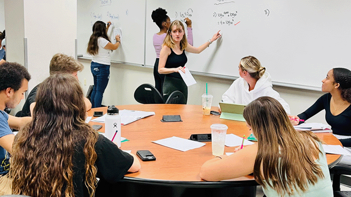A blonde LA in a black tank top gesturing to a white board behind a table of engaged students