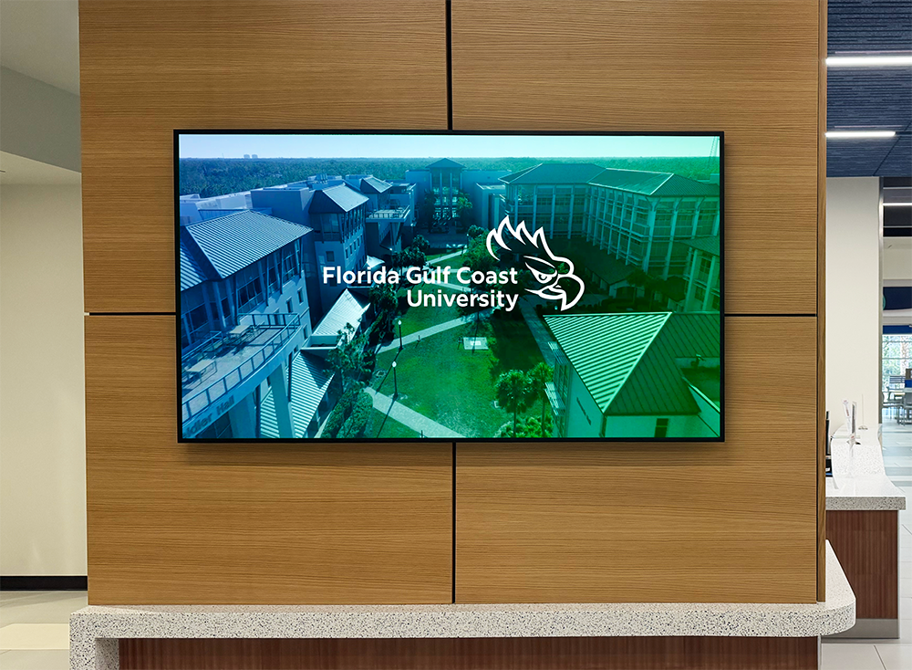 Digital display mounted on a wooden wall showing an aerial view of the Florida Gulf Coast University campus with green landscaping, metal‑roofed buildings, and the FGCU logo with an eagle head overlaid on the image.
