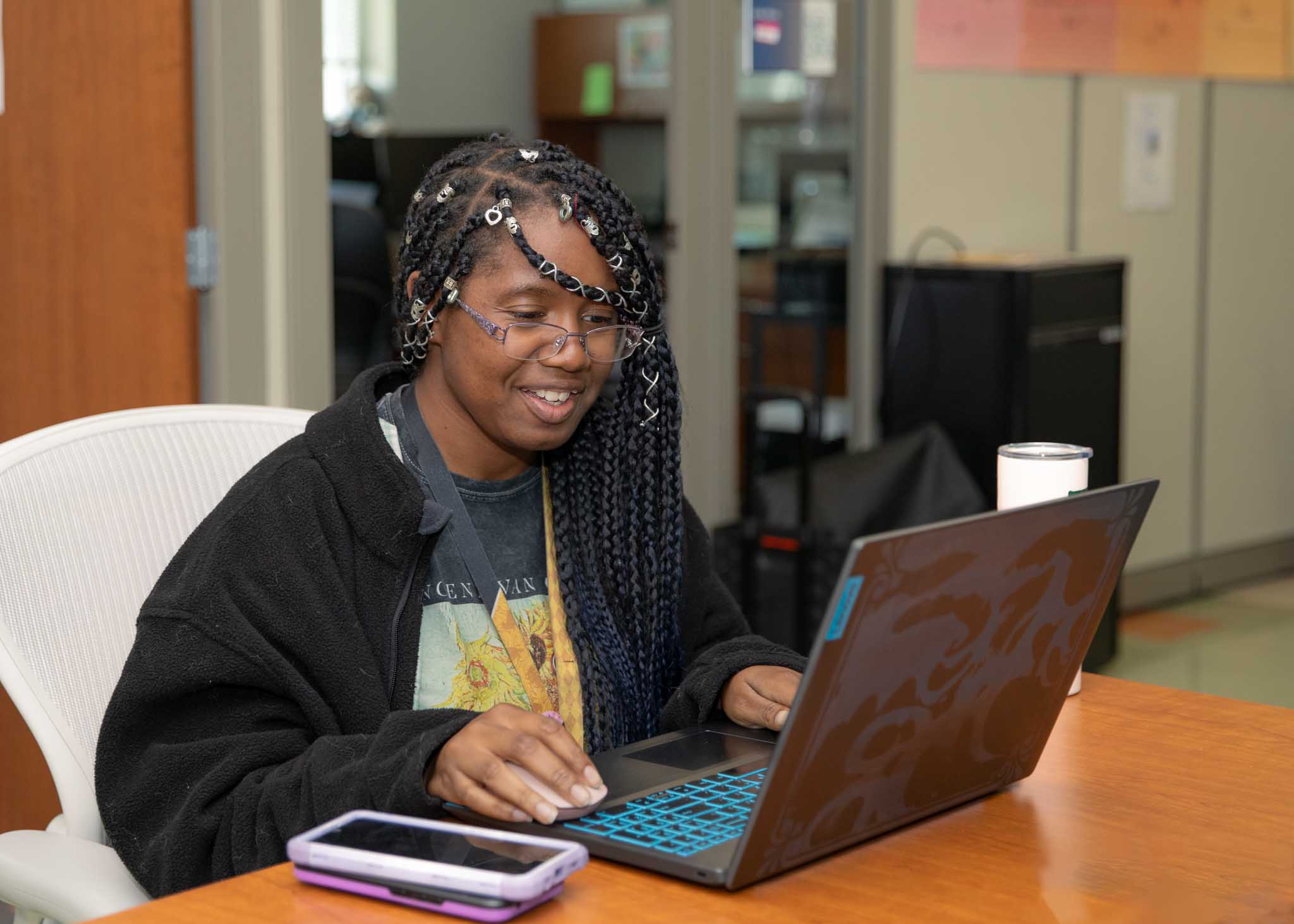 Female student in a black jacket working on a laptop and smiling.