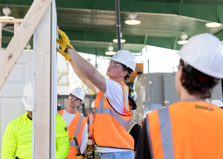 A student wearing PPE measures a wooden frame for a building