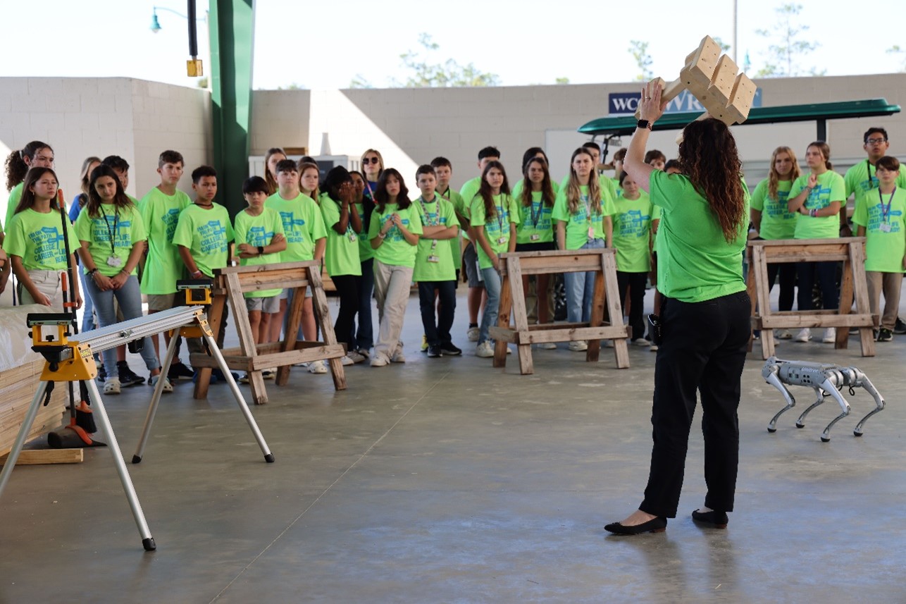Woman holds up a wooden stool to a crowd of seventh graders dressed in neon green shirts