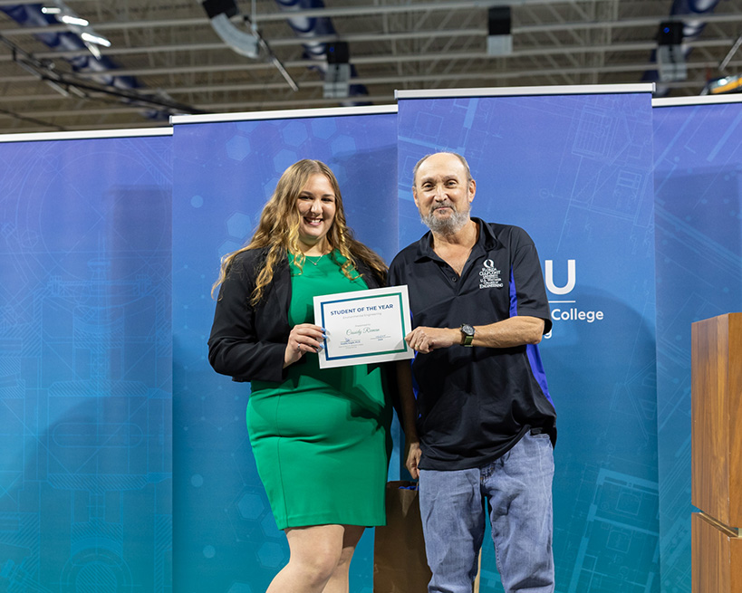 A woman in a blazer and green dress holds a certificate with a man in a black polo