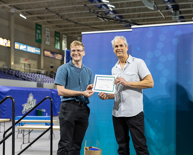 A man in glasses and a blue button down holds a certificate and stands near a man in a light button down