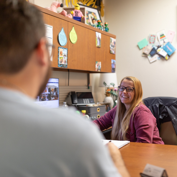 An advisor sits at her desk smiling at a student