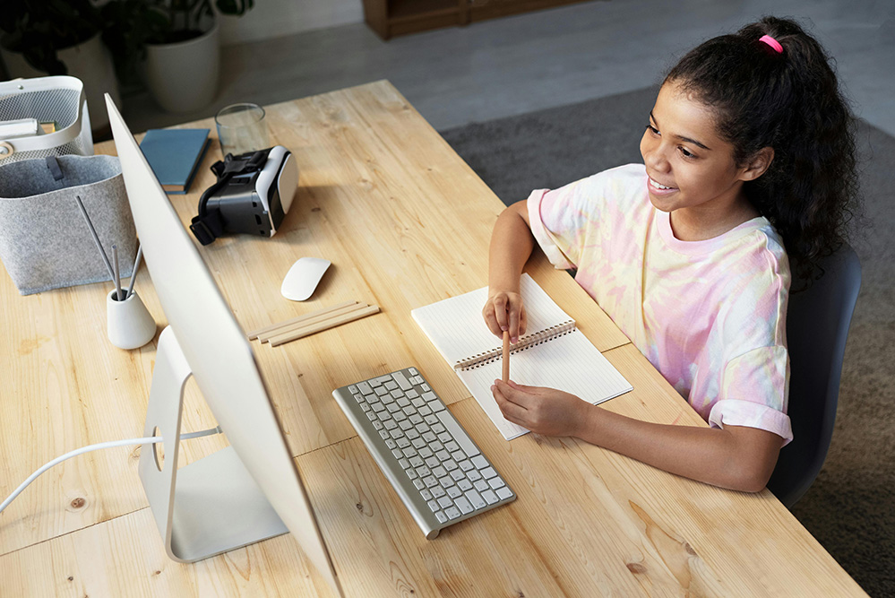A young girl sits at a desk looking at a computer and smiling
