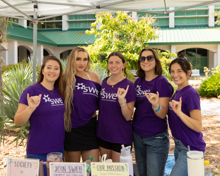 Five students wearing purple shirts that say SWE do "wings up" with their hands