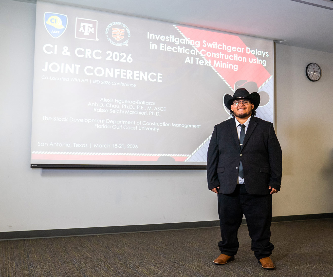 Alexis wears a large black hat and stands in front of a screen with his conference presentation
