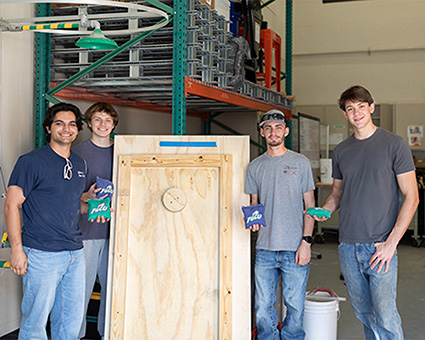 Four students in t-shirts and jeans stand near a wooden cornhole form while holding bean bags