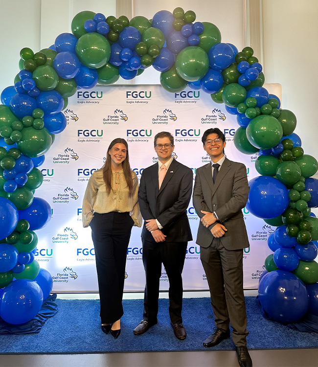 Three students stand in front of a blue and green balloon arch and an FGCU backdrop