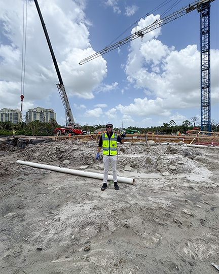 A man in a yellow construction vest, hard hat, and gloves stands on a construction site