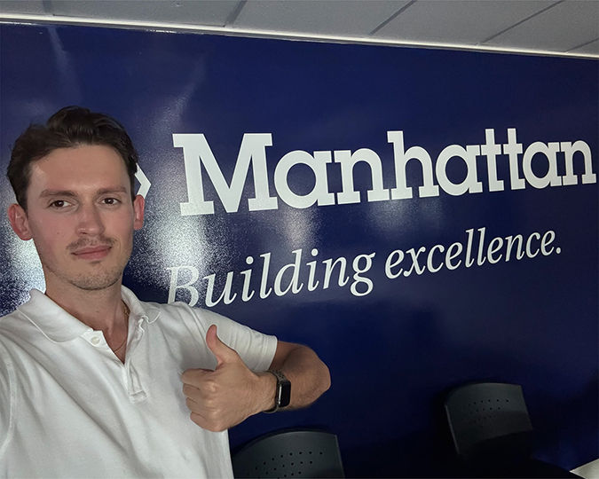 A man in a white polo holds a thumbs up in front of a sign that says Manhattan Building Excellence