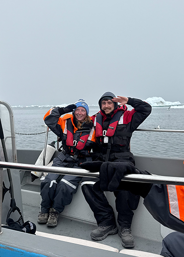 Two people in heavy coats and life jackets sit in a boat, smiling