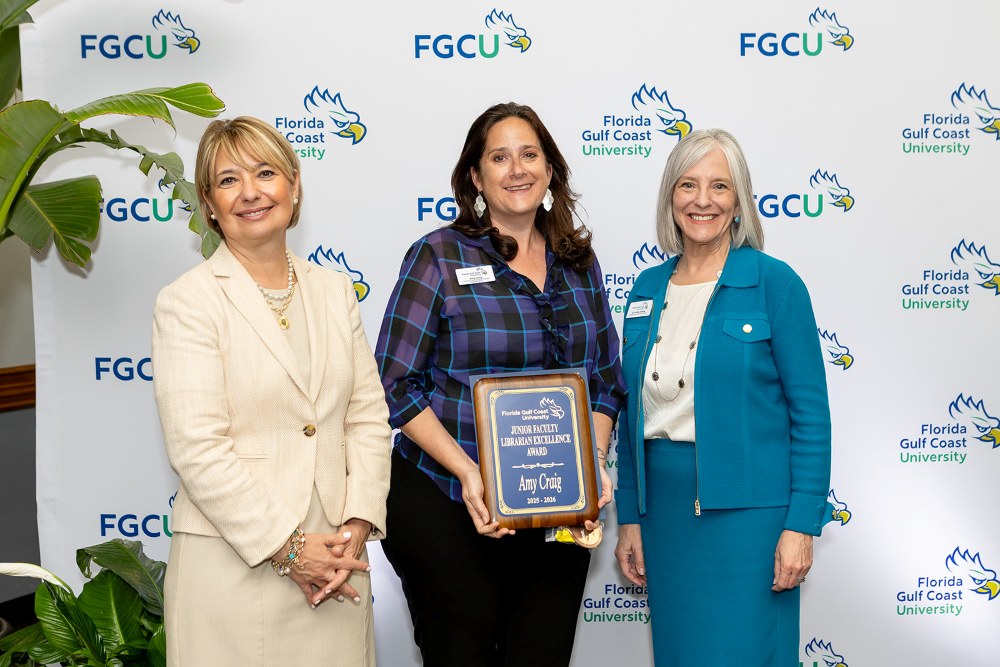 Three women stand in front of a Florida Gulf Coast University backdrop at the 2026 Celebration of Excellence Award Ceremony. The woman in the center holds a plaque recognizing the Junior Librarianship Excellence Award, while the two women beside her stand smiling.