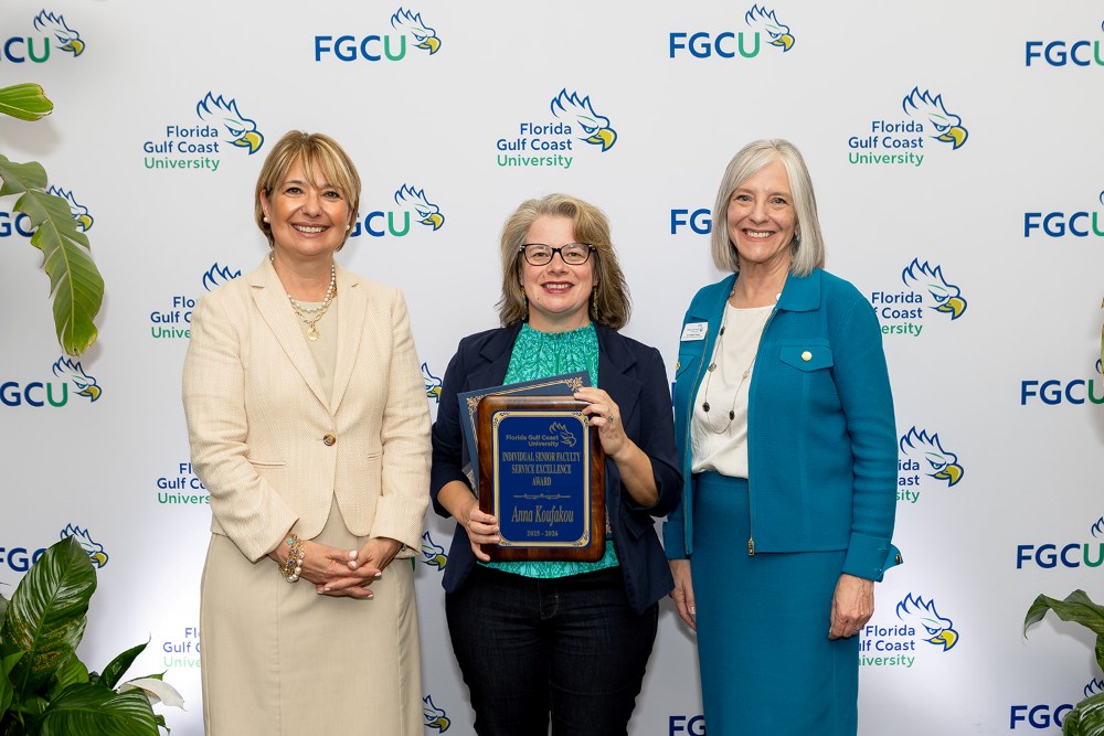 Three women stand in front of a Florida Gulf Coast University backdrop at the 2026 Celebration of Excellence Award Ceremony. The woman in the center holds a plaque recognizing the Individual Faculty Service Excellence Award, while the two women beside her stand smiling.