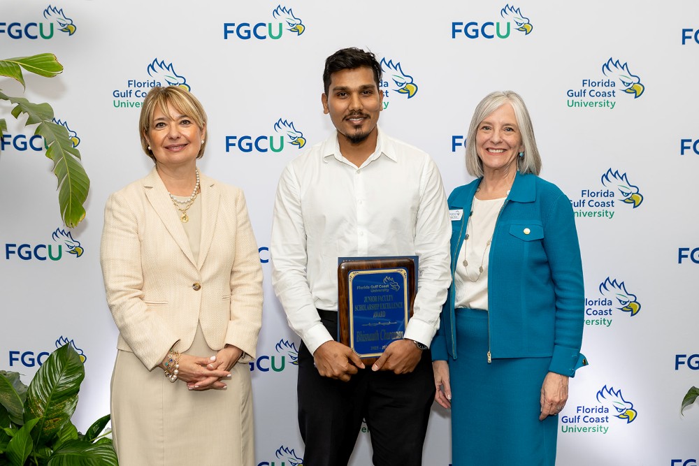 Two women and one man stand in front of a Florida Gulf Coast University backdrop at the 2026 Celebration of Excellence Award Ceremony. The man in the center holds a plaque recognizing the Junior Faculty Scholarship Excellence Award, while the two women beside him stand smiling.