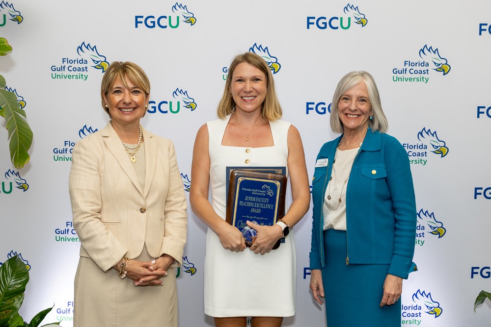 Three women stand in front of a Florida Gulf Coast University backdrop at the 2026 Celebration of Excellence Award Ceremony. The woman in the center holds a plaque recognizing the Junior Faculty Teaching Excellence Award, while the two women beside her stand smiling.