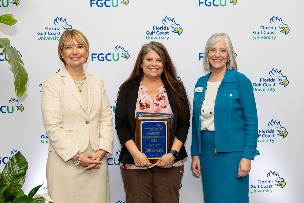Three women stand in front of a Florida Gulf Coast University backdrop at the 2026 Celebration of Excellence Award Ceremony. The woman in the center holds a plaque recognizing the Junior Faculty Scholarship Excellence Award, while the two women beside her stand smiling.