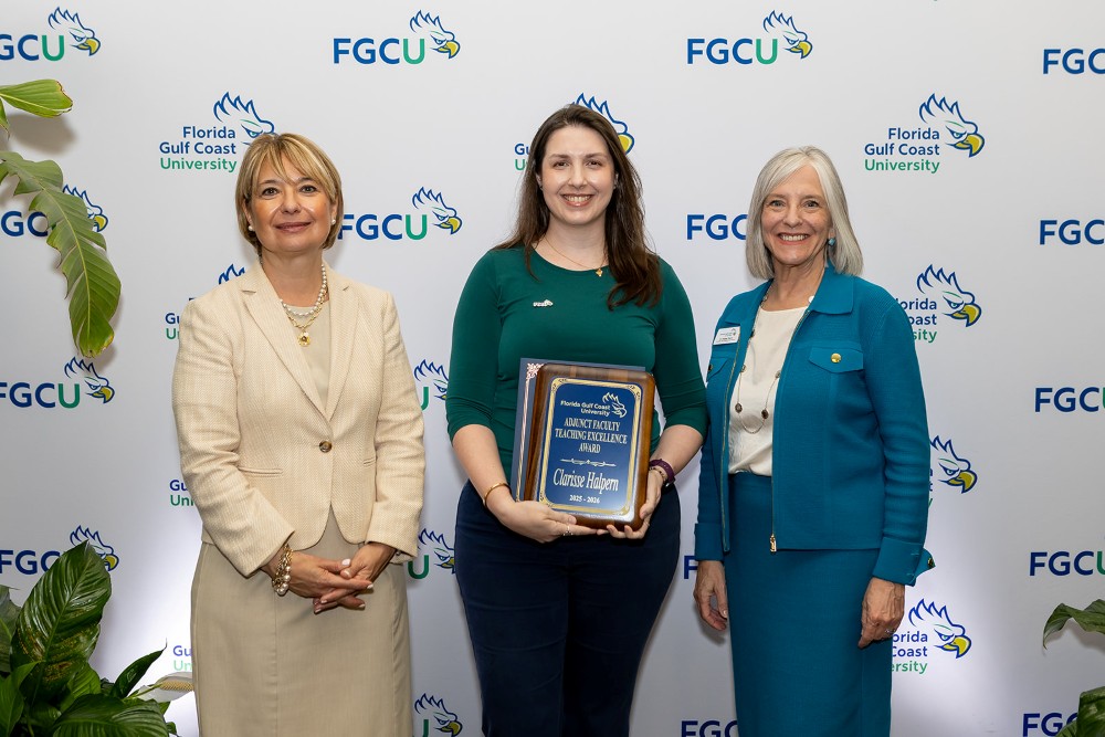 Three women stand in front of a Florida Gulf Coast University backdrop at the 2026 Celebration of Excellence Award Ceremony. The woman in the center holds a plaque recognizing the Adjunct Faculty Teaching Excellence Award, while the two women beside her stand smiling.