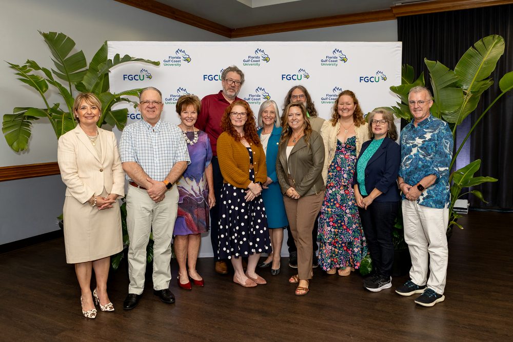 Group photo of eleven individuals standing indoors in front of a white Florida Gulf Coast University (FGCU) branded backdrop, flanked by large green plants. The individuals are arranged in two rows and dressed in business and business-casual attire. This group was recognized with the Team Service Excellence Award at the 2026 Celebration of Excellence ceremony at Florida Gulf Coast University.