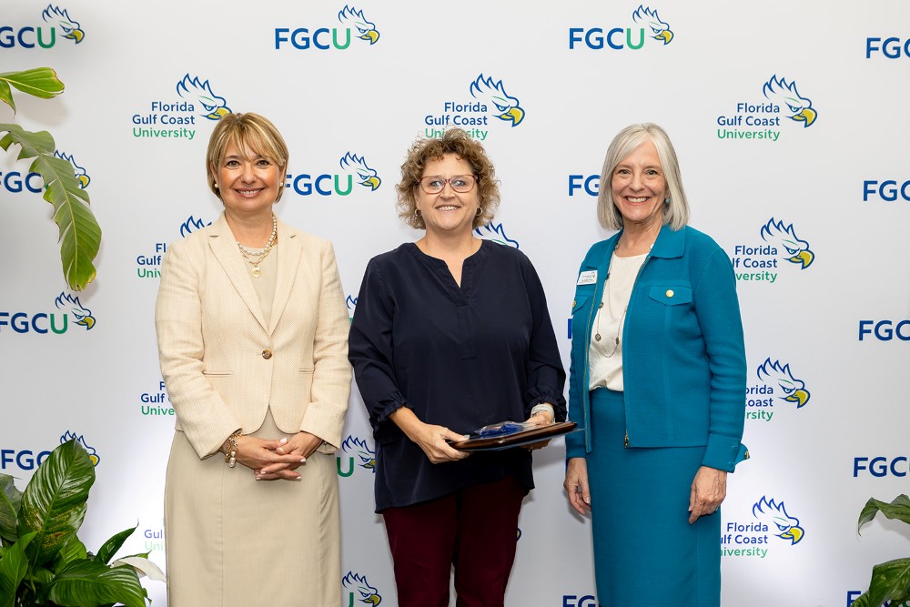 Three women stand in front of a Florida Gulf Coast University backdrop at the 2026 Celebration of Excellence Award Ceremony. The woman in the center holds a plaque recognizing the Senior Instructor Teaching Excellence Award, while the two women beside her stand smiling.