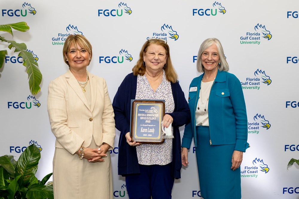 Three women stand in front of a Florida Gulf Coast University backdrop at the 2026 Celebration of Excellence Award Ceremony. The woman in the center holds a plaque recognizing the Individual Faculty Service Excellence Award, while the two women beside her stand smiling.