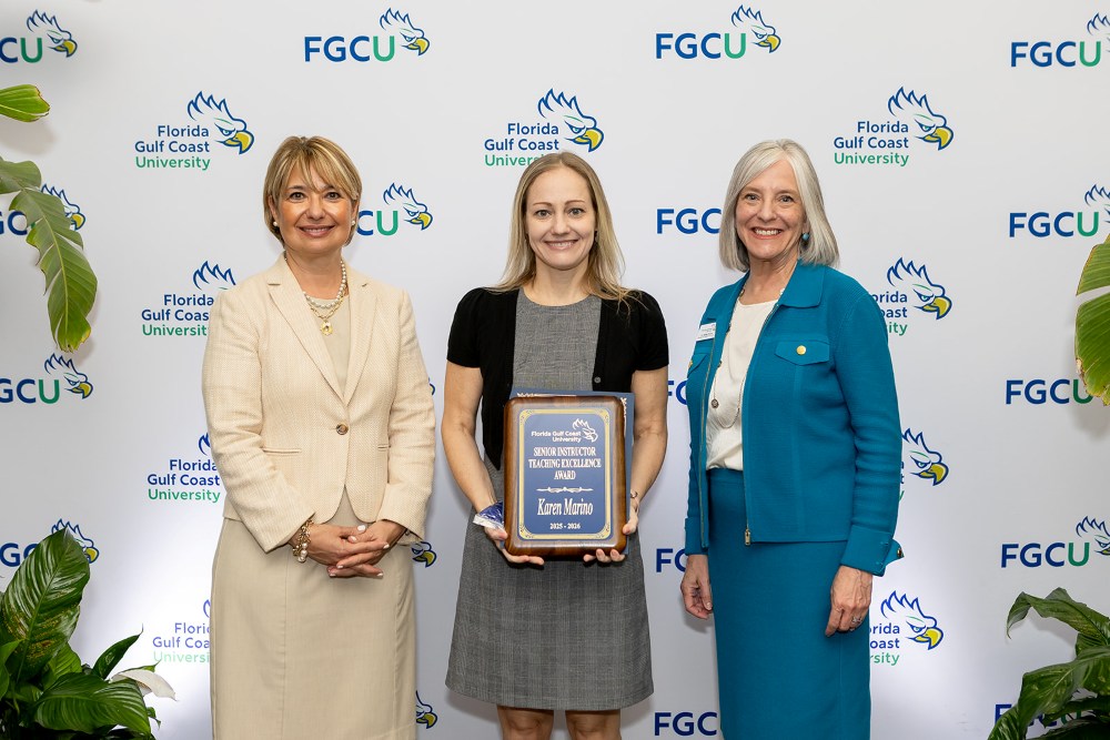 Three women stand in front of a Florida Gulf Coast University backdrop at the 2026 Celebration of Excellence Award Ceremony. The woman in the center holds a plaque recognizing the Senior Instructor Teaching Excellence Award, while the two women beside her stand smiling.