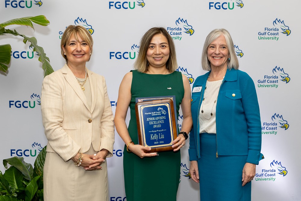 Three women stand in front of a Florida Gulf Coast University backdrop at the 2026 Celebration of Excellence Award Ceremony. The woman in the center holds a plaque recognizing the Junior Advising Excellence Award, while the two women beside her stand smiling.