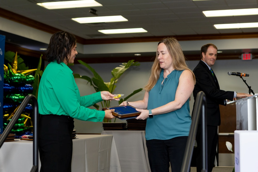 Award recipient accepts a plaque during the 2026 Celebration of Excellence ceremony at Florida Gulf Coast University, with an announcer at a podium nearby.