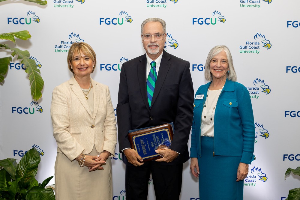 Two women and one man stand in front of a Florida Gulf Coast University backdrop at the 2026 Celebration of Excellence Award Ceremony. The man in the center holds a plaque recognizing the Adjunct Faculty Teaching Excellence Award, while the two women beside him stand smiling.