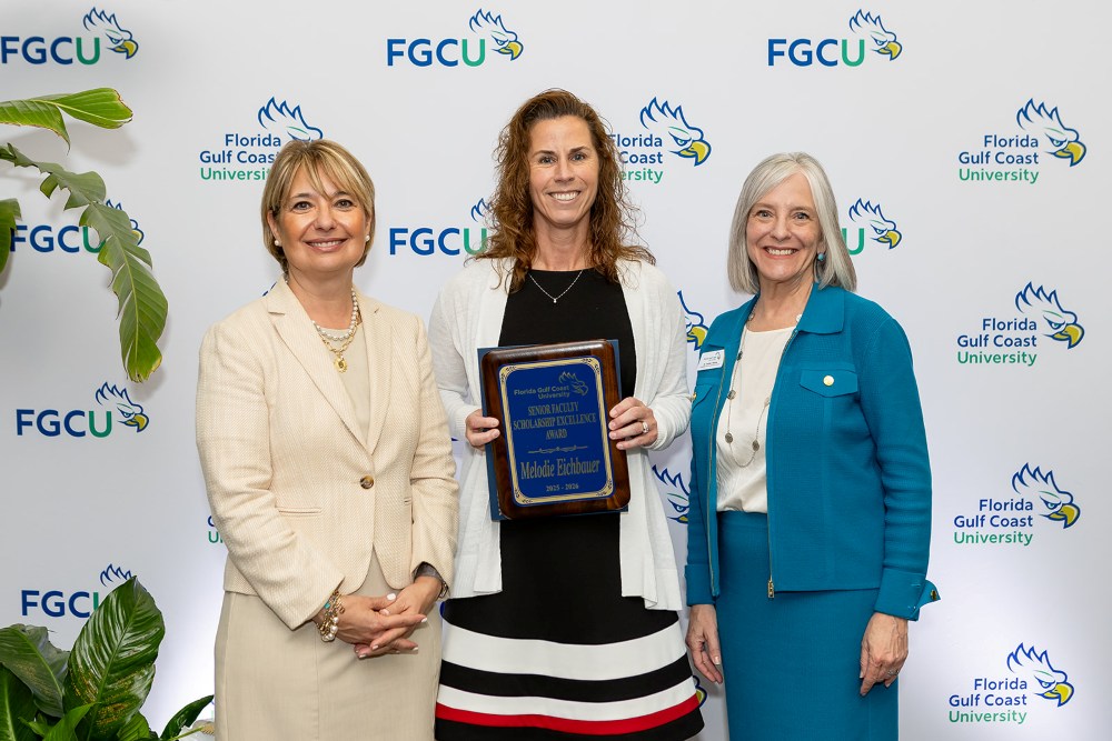 Three women stand in front of a Florida Gulf Coast University backdrop at the 2026 Celebration of Excellence Award Ceremony. The woman in the center holds a plaque recognizing the Senior Faculty Scholarship Excellence Award, while the two women beside her stand smiling.