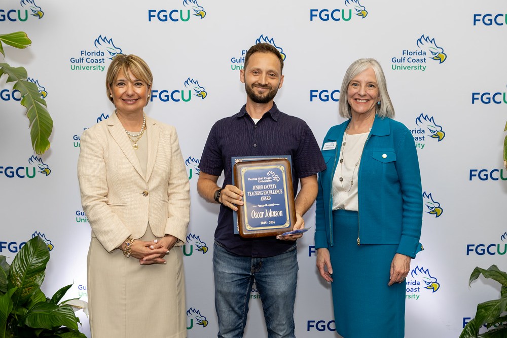 Two women and one man stand in front of a Florida Gulf Coast University backdrop at the 2026 Celebration of Excellence Award Ceremony. The man in the center holds a plaque recognizing the Junior Faculty Teaching Excellence Award, while the two women beside him stand smiling.