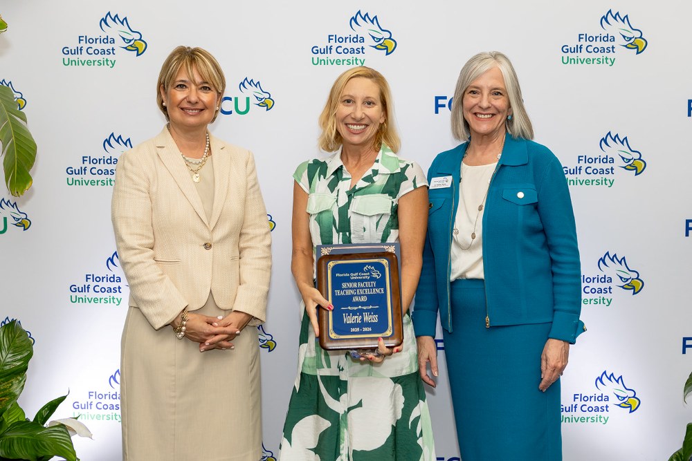 Three women stand in front of a Florida Gulf Coast University backdrop at the 2026 Celebration of Excellence Award Ceremony. The woman in the center holds a plaque recognizing the Senior Faculty Teaching Excellence Award, while the two women beside her stand smiling.
