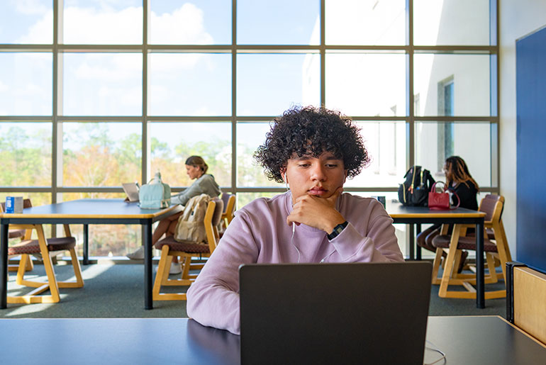 Student in a light purple sweatshirt working on a laptop in a bright, modern study area.