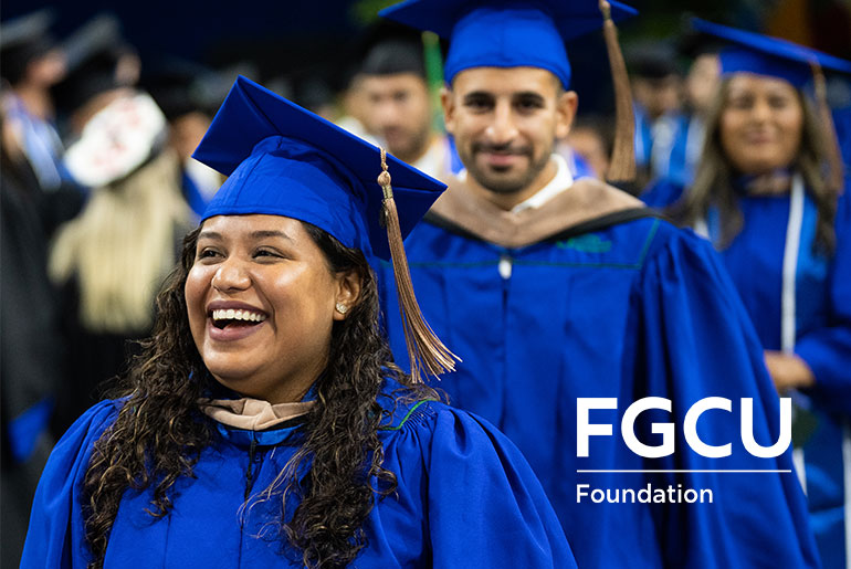 A group of graduates in blue caps and gowns smile and celebrate during a commencement ceremony, with “FGCU Foundation” displayed in the corner.