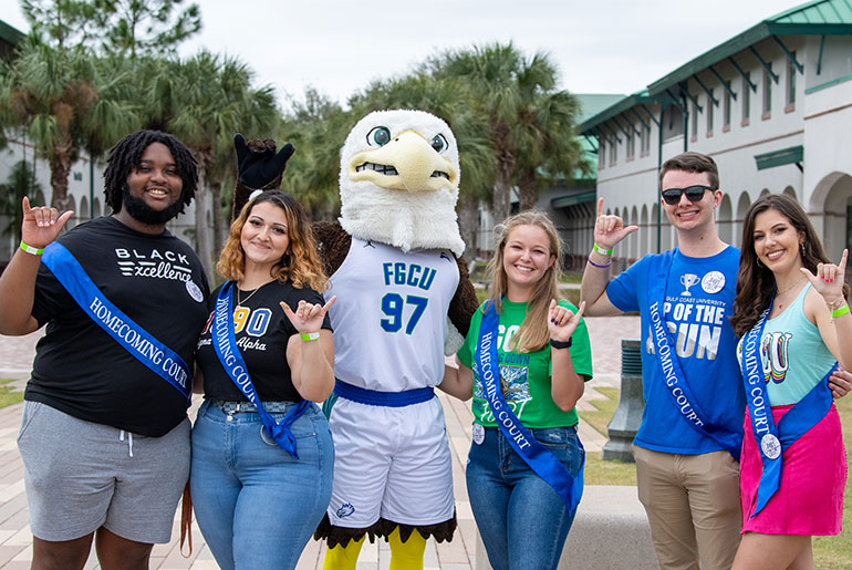  group of five college students stand outdoors on a campus walkway alongside an eagle mascot wearing an FGCU jersey. The students wear casual clothing with blue “Homecoming Court” sashes and make hand gestures, smiling and posing together. Palm trees and campus buildings are visible in the background.