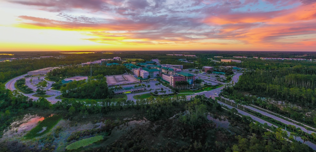Aerial view of Florida Gulf Coast University surrounded by lush greenery and forests. Buildings are arranged in a circular pattern, connected by roads. The sky is illuminated with vibrant hues of orange, pink and purple from a rising sun. 
