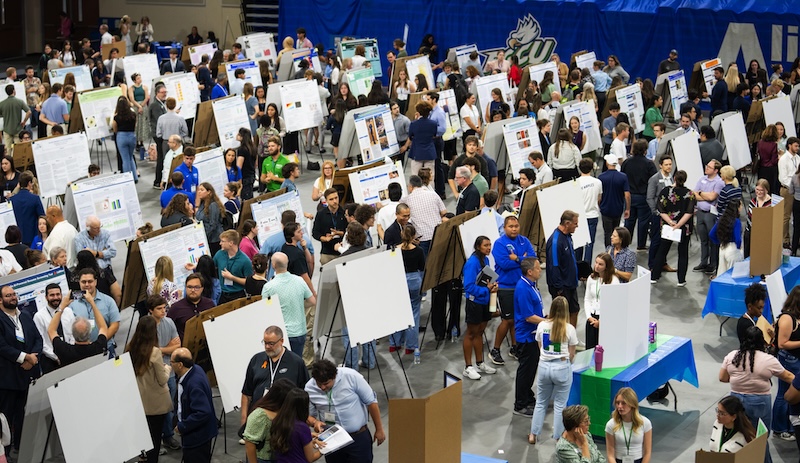 A large crowd fills Alico Arena for a research showcase, with rows of poster boards displayed across the floor. Students, faculty and visitors walk among the exhibits, discussing projects and presenting their work. A blue FGCU banner hangs in the background.  