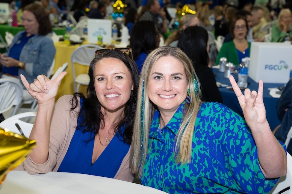 Two women are sitting next to each other in front of a white table. The woman to the left is wearing a blue blouse with a brown cardigan. The woman to the right is wearing a blue and green dress and has blue and green tinsel in her hair. Both women are doing the Wings Up sign. In the background are other tables with people sitting down eating lunch.  
