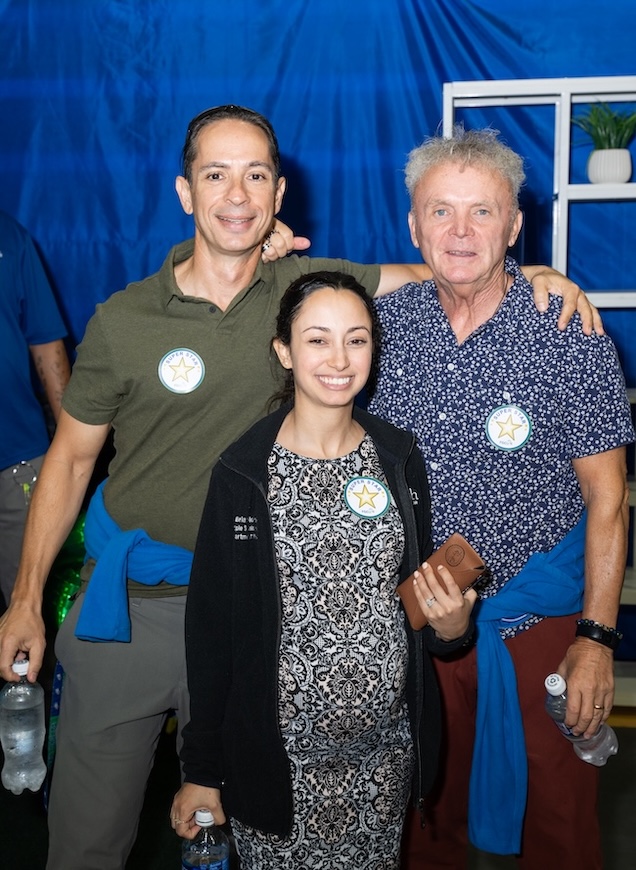 hree FGCU employees pose for a group photo at the Fall Welcome Celebration. The man on the left is wearing a dark green polo and grey pants. The man to the right is wearing a blue and white button-up shirt and maroon pants. The woman in the middle is wearing a black and white dress and a black zip-up sweater. All three are standing in front of a blue wall and are wearing star stickers on their clothing. 
