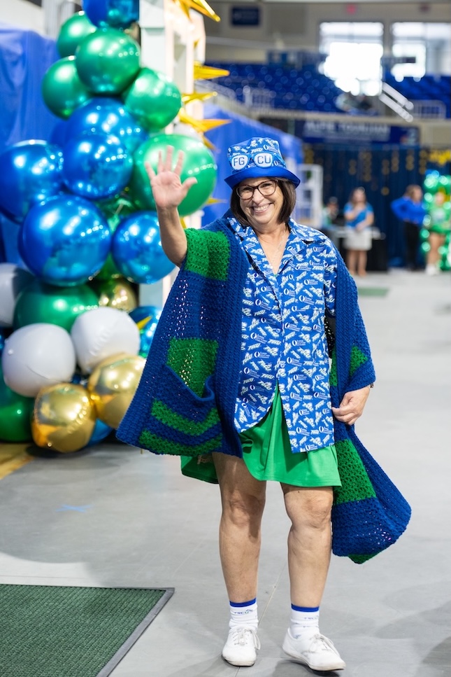 Kimberly Hobart walks up to the stage dressed in a green skirt, a FGCU logo blouse and hat, a green and blue knitted scarf, and white sneakers. A blue, green and gold balloon arch and blue bleachers are in the background. 