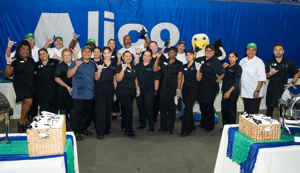 A group of FGCU Dining Services staff are wearing black polos and doing the “Wings Up” sign as they pose for a group photo with Azul, the FGCU mascot. They are standing in front of a backdrop with the words ‘Alico’ in white.  