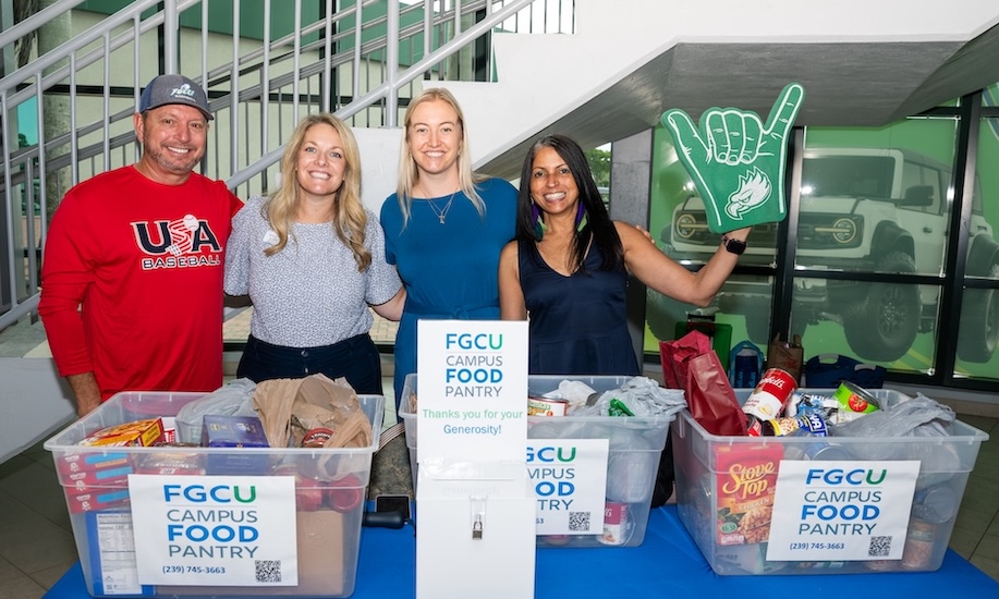 Four FGCU employees are standing behind a table with three clear plastic containers filled with canned goods and non-and perishable items for the FGCU campus Food Pantry. The person on the far left is wearing a grey FGCU baseball cap and a red long sleeve shirt. The second person is wearing a black and white blouse and black pants. The next person is wearing a blue dress and the person to the far right is wearing a dark blue blouse and is holding up a green FGCU foam finger. White stairs can be seen in the background.  