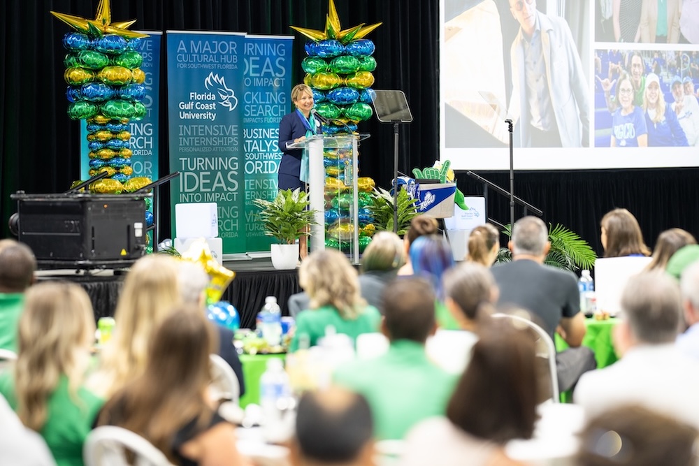 Dressed in a dark blue blazer and skirt suit, and a green and blue scarf, President Aysegul Timur is addressing the crowd at Alico Arena from the stage. To the right and left side of her are green, blue and gold balloon towers. Behind her are blue and green banners with the words like 'Turning Ideas into Impact' and 'Intensive Internships’ in white.  