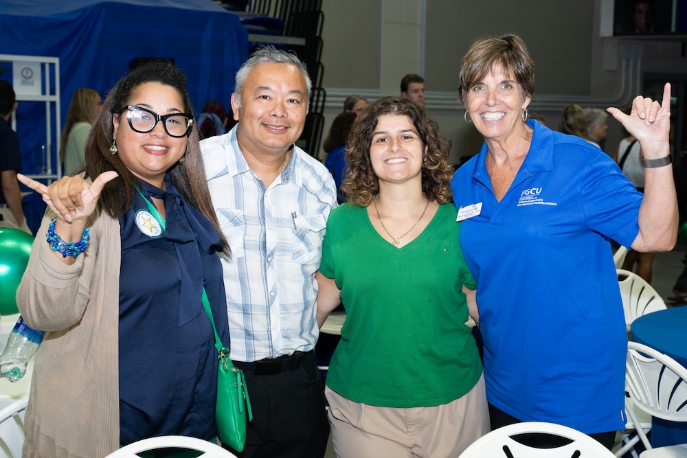 Four FGCU employees pose together for a photo. From left to right: one employee wears a dark blue blouse with a beige cardigan, carries a green purse and is doing the Wings Up sign. The next person is wearing a light blue plaid shirt and black pants. The third person wears a green blouse with beige pants, and the fourth is in a blue FGCU polo shirt while doing the Wings Up sign. 