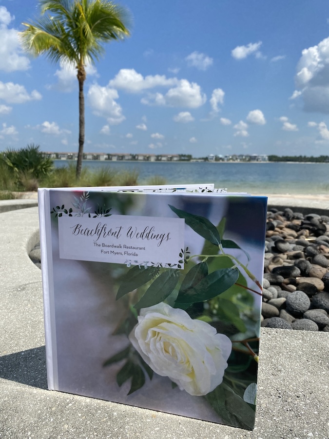 A Beachfront Wedding book is displayed with FGCU’s waterfront Lake Como in the background as well as a palm tree on the left side and a few homes in the background.  The wedding book has a white rose on the front cover. The book is standing on top of stone that is part of a fire fit with grey and brown pebbles.  