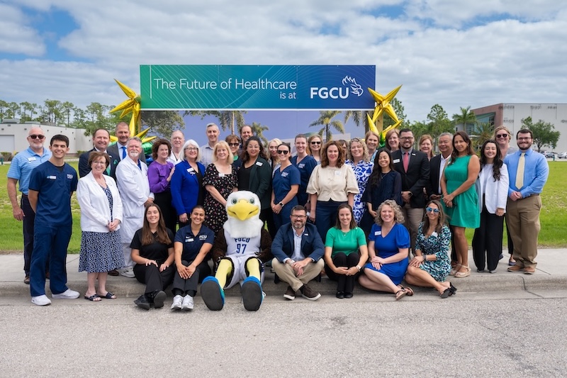 Faculty and staff from the Marieb College of Health & Human Services and FGCU’s mascot Azul pose for a group photo outside in front of a poster with a rendering of academic building 10.