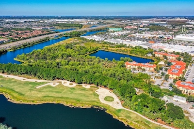 Aerial view of the future location of FGCU’s workforce housing. A landscaped golf course bordered by a lake with dense green trees and vegetation can be seen south of the housing site. FGCU residential student housing, West Lake Village, is to the east of the site. Multiple waterways and ponds are visible, along with a clear blue sky. 